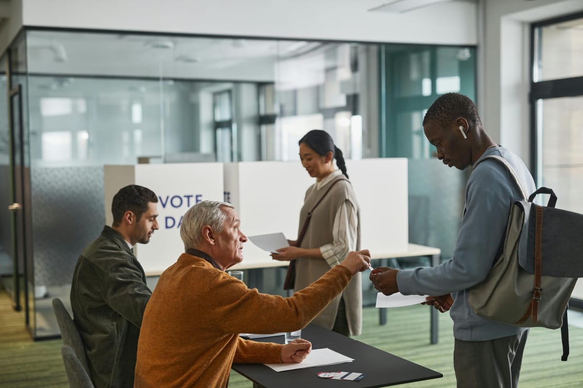 Photo: The interior of a polling site. Two poll workers are seated at tables and are interacting with two voters. 